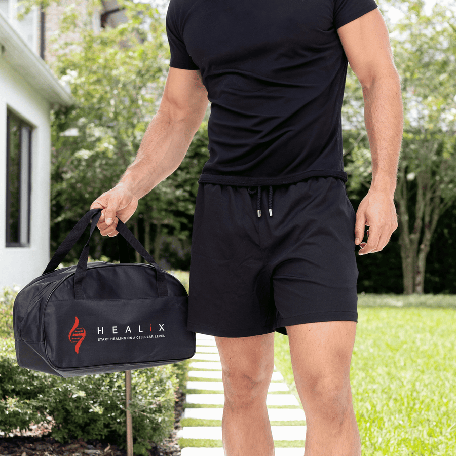 A man in a black t-shirt and shorts stands outside on a sunny day, holding the HEALiX Infrared HEALiX Flow gym bag with the red and white logo and DNA motif. Lush green grass and trees can be seen behind him.