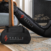 A black HEALiX Flow bag and a HEALiX Flow leg compression sleeve by HEALiX Infrared rest on a patterned rug near a chair and fireplace, both displaying the HEALiX Infrared logo and slogan, "Start healing on a cellular level.