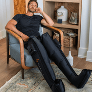A man relaxes in a modern living room, eyes closed, wearing HEALiX Infrared’s HEALiX Flow black leg compression sleeves with control panels, surrounded by wooden shelves and neutral decor.