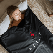 A woman relaxes on a cushion under a HEALiX-Z Sauna Blanket by HEALiX Infrared, enjoying the comfort of this premium infrared sauna blanket while lying on a woven mat.