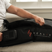 A person in a white shirt sits on a couch, zipping up the black HEALiX Infrared HEALiX Flow device with its digital control panel. The logo and the phrase "Start healing on a cellular level" are visible.