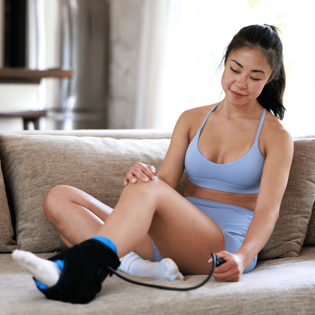 A woman sits on a beige couch, smiling as she uses the Icebound Essentials Frost Fit Compression Ankle for hot/cold therapy. She holds the strap for targeted, effective ankle recovery and appears relaxed and comfortable.