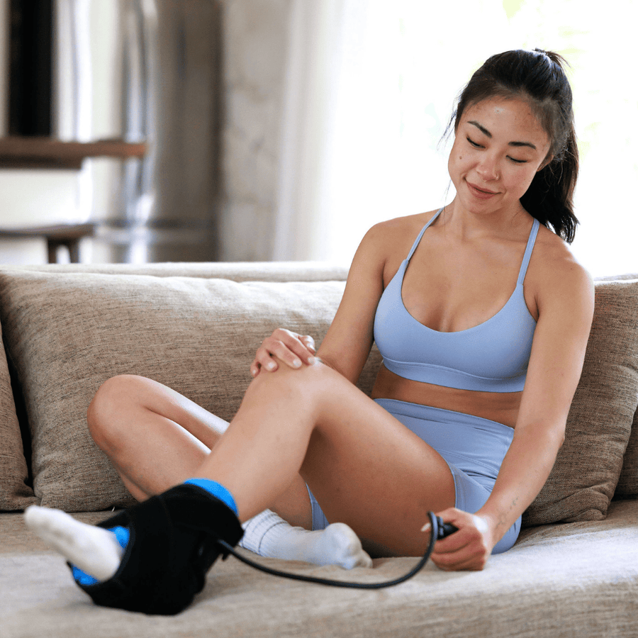 A woman sits on a beige couch, smiling as she uses the Icebound Essentials Frost Fit Compression Ankle for hot/cold therapy. She holds the strap for targeted, effective ankle recovery and appears relaxed and comfortable.