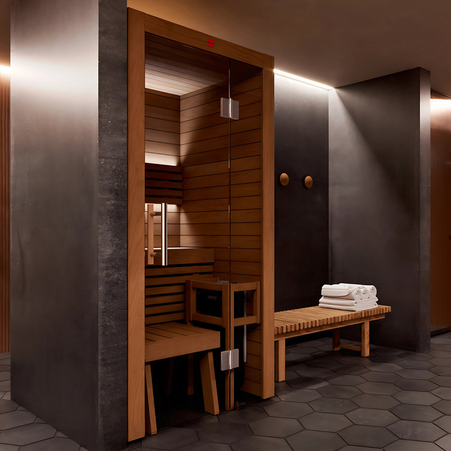 The LuxeWell Life Harvia Utu indoor sauna features a heat-treated Nordic spruce interior, glass door, benches, and premium heating unit. A wooden bench with towels sits outside against dark walls and hexagonal tiles under soft lighting.