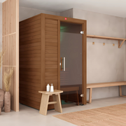 The LuxeWell Life Harvia Utu, a heat-treated 1-person indoor sauna with a glass door, is set in a minimalist room with a small wooden bench holding two bottles and an adjacent wall-mounted coat rack and bench.
