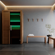 A modern, minimalist spa room features the LuxeWell Life Harvia Utu heat-treated 1-person indoor sauna, a bench with folded towels, wall hooks, a small table, and a potted plant against concrete and tile. Soft lighting enhances the calm ambiance.