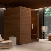 A LuxeWell Life Harvia Utu heat-treated 1-person indoor sauna is set in a minimalist, well-lit room with light floors, a lounge chair, a small table, and a large window overlooking lush green trees.