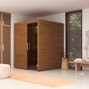The LuxeWell Life Harvia Utu is a modern, heat-treated Nordic spruce indoor sauna for one person with a glass door, set in a bright minimalist room featuring a wooden bench with towels, plant, hanging robe, and woven rug on white tile.
