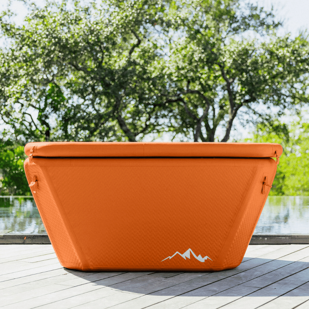 The Big Tex Endurance Plunge Tub by Icebound Essentials, an orange trapezoid-shaped inflatable tub with a white mountain logo, sits on a wooden deck near green trees and a reflective body of water.