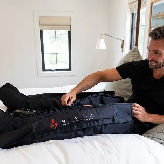A man in a black shirt sits on a bed, using the HEALiX Infrared HEALiX Flow Pro Compression Boot for pneumatic therapy, with natural light coming through the window as part of his recovery routine.
