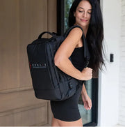 A woman in a black dress stands by a door, smiling with a large black backpack featuring the HEALiX Infrared logo—ready for recovery with the HEALiX Flow Pro Compression Boot inside.
