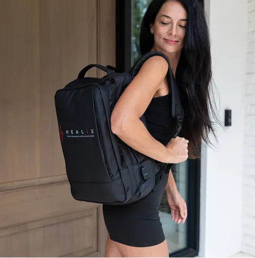A woman in a black dress stands by a door, smiling with a large black backpack featuring the HEALiX Infrared logo—ready for recovery with the HEALiX Flow Pro Compression Boot inside.