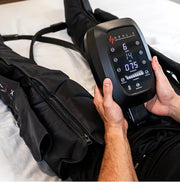 A person uses a HEALiX Flow Pro Compression Boot by HEALiX Infrared while lying on a bed, with the display showing recovery settings to boost circulation, much like other compression devices.