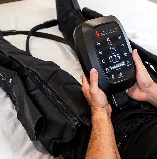 A person uses a HEALiX Flow Pro Compression Boot by HEALiX Infrared while lying on a bed, with the display showing recovery settings to boost circulation, much like other compression devices.