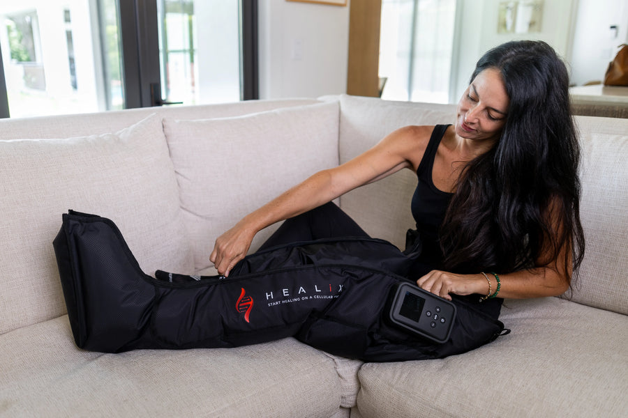 A woman sits on a beige couch, smiling as she places her leg into the black HEALiX Infrared HEALiX Flow compression therapy device with its control panel.