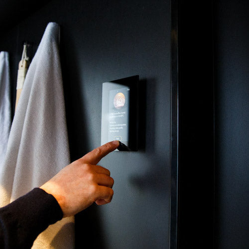 A hand reaches for a Harvia Fenix WiFi Remote Control Panel by Harvia, mounted on a dark wall, with a white towel hanging in the background.