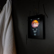 The Harvia Fenix WiFi Remote Control Panel by Harvia, mounted on a dark wall with water droplets, shows 70°F; a white towel nearby enhances the atmosphere of relaxation and advanced comfort.