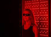 A woman wearing Mito Red Light | Red Light Therapy Glasses IR5 (Extra Dark) by Mito Red Light stands before a panel of bright red LEDs, demonstrating the importance of eye protection during red and near-infrared light treatments.