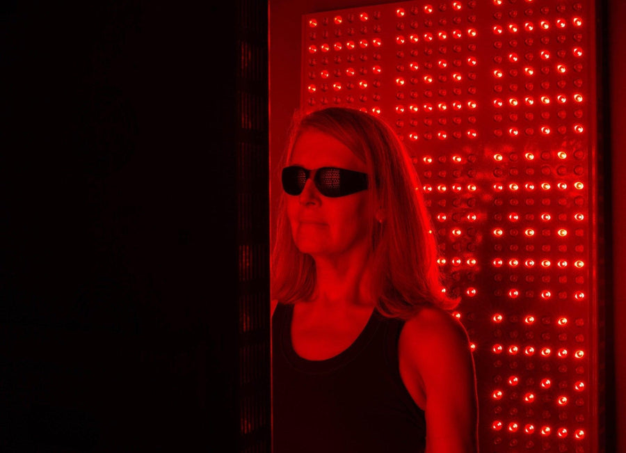 A woman wearing Mito Red Light | Red Light Therapy Glasses IR5 (Extra Dark) by Mito Red Light stands before a panel of bright red LEDs, demonstrating the importance of eye protection during red and near-infrared light treatments.