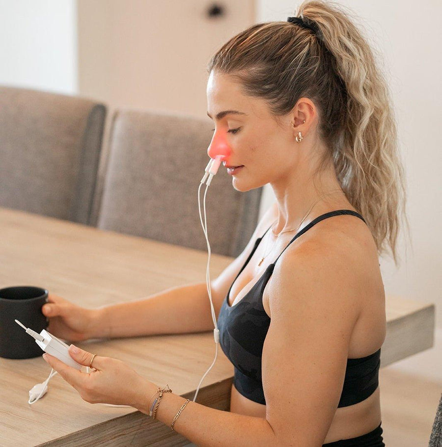 A woman in athletic wear sits at a table holding a mug and the MitoBOOST™ Intranasal Device System by Mito Red Light, enjoying its red light therapy for sinus health, immune support, and brain health.