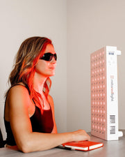 A woman in black sunglasses sits at a desk with a notebook, facing the MitoMID 2.0 Red Light Therapy Panel by Mito Red Light, its powerful LED glow illuminating her face and upper body.