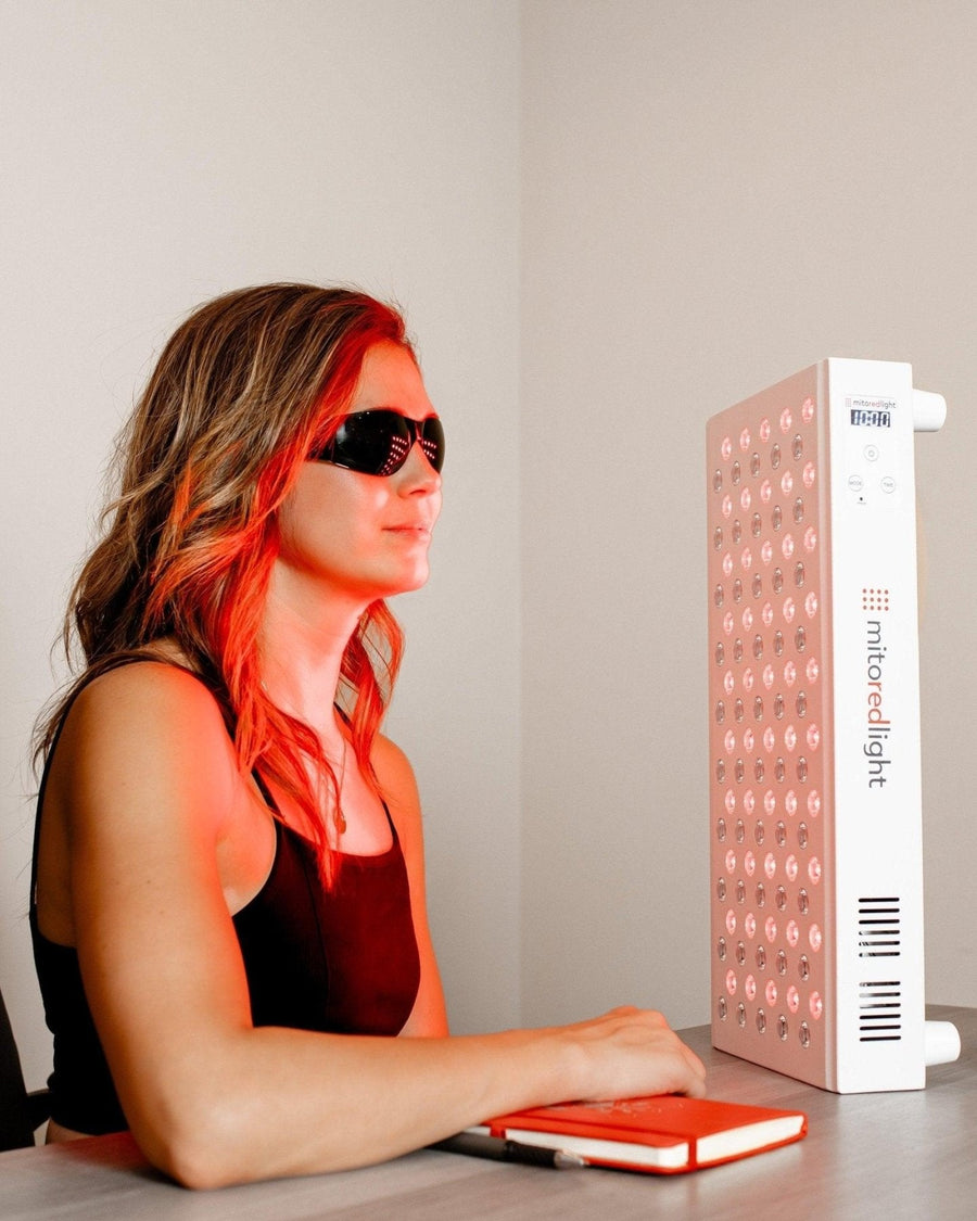 A woman in black sunglasses sits at a desk with a notebook, facing the MitoMID 2.0 Red Light Therapy Panel by Mito Red Light, its powerful LED glow illuminating her face and upper body.