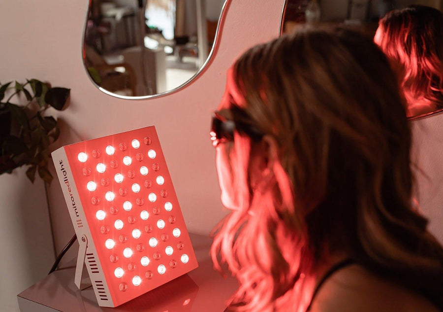 A person wearing protective glasses sits in front of a Mito Red Light MitoMIN 2.0 | Red Light Therapy Panel, which is turned on and emitting red light in a home setting with a mirror and plant visible in the background.