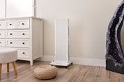 A tall, white air purifier stands on wood flooring in a corner next to a white dresser, with a pouf, stool, part of a geode, and the MitoPRO+ | Red Light Therapy Panel Series v2 by Mito Red Light visible nearby.