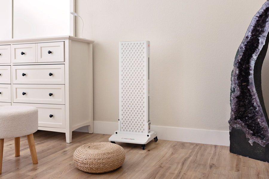 A tall, white air purifier stands on wood flooring in a corner next to a white dresser, with a pouf, stool, part of a geode, and the MitoPRO+ | Red Light Therapy Panel Series v2 by Mito Red Light visible nearby.