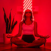 A woman in a white tank top and dark shorts sits cross-legged, meditating with protective glasses before the Mito Red Light MitoPRO+ | Red Light Therapy Panel Series v2, featuring quad wavelength red and near-infrared light. A potted plant sits nearby.