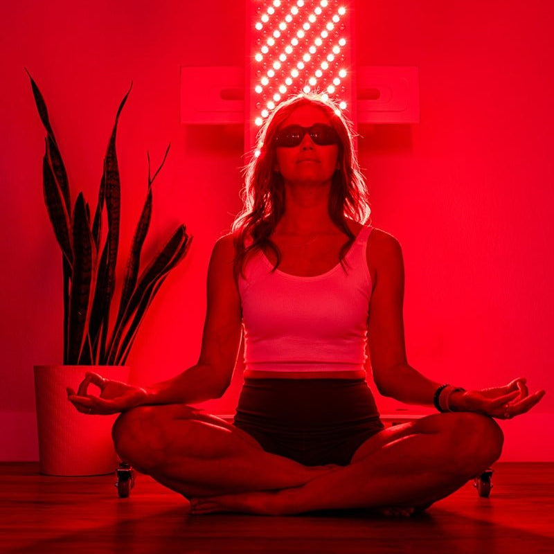 A woman in a white tank top and dark shorts sits cross-legged, meditating with protective glasses before the Mito Red Light MitoPRO+ | Red Light Therapy Panel Series v2, featuring quad wavelength red and near-infrared light. A potted plant sits nearby.