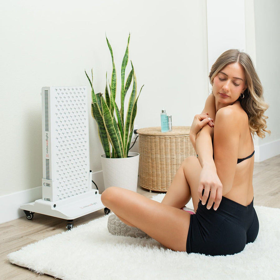 A woman in black shorts and a bra sits on a wooden floor, applying lotion to her arm next to the Mito Red Light MitoPRO+ | Red Light Therapy Panel Series v2. A green potted plant and wicker basket are visible in the background.