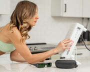 A woman in a light green sports bra adjusts the MitoPRO+ | Red Light Therapy Panel Series v2 by Mito Red Light in a bright kitchen, with protective glasses and a black case beside her on the counter.