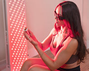 A woman in sunglasses and athletic wear sits beside a tall Mito Red Light MitoPRO+ | Red Light Therapy Panel Series v2, the red and near infrared light illuminating her as she examines a small container.