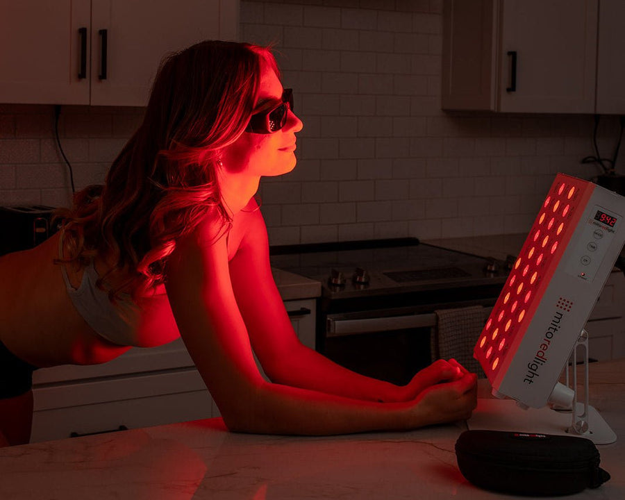 A woman in sunglasses leans on a kitchen counter, illuminated by the vivid red glow of the Mito Red Light MitoPRO+ | Red Light Therapy Panel Series v2 with quad wavelength design.