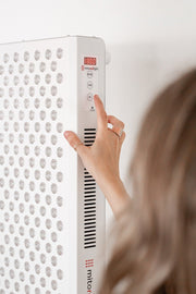 A person with light brown hair presses a button on a white MitoPRO+ | Red Light Therapy Panel Series v2 by Mito Red Light, featuring multiple small LEDs and a digital display reading “1880.”.