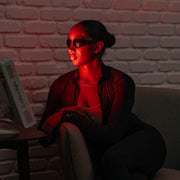 A woman in black sunglasses and jacket sits on a chair, facing the Mito Red Light MitoPRO+ | Red Light Therapy Panel Series v2 with quad wavelength design. The device emits red light; a white brick wall is in the background.
