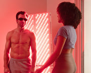 A shirtless man in sunglasses stands before the Mito Red Light MitoPRO+ | Red Light Therapy Panel Series v2, featuring quad wavelength red & near infrared light, while a woman in activewear faces him, both bathed in its therapeutic red glow.