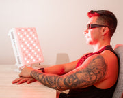 A person with sunglasses and a tattooed arm sits at a table using the MitoPRO+ Red Light Therapy Panel Series v2 by Mito Red Light, which emits bright red light onto their face and upper body with its quad wavelength design.