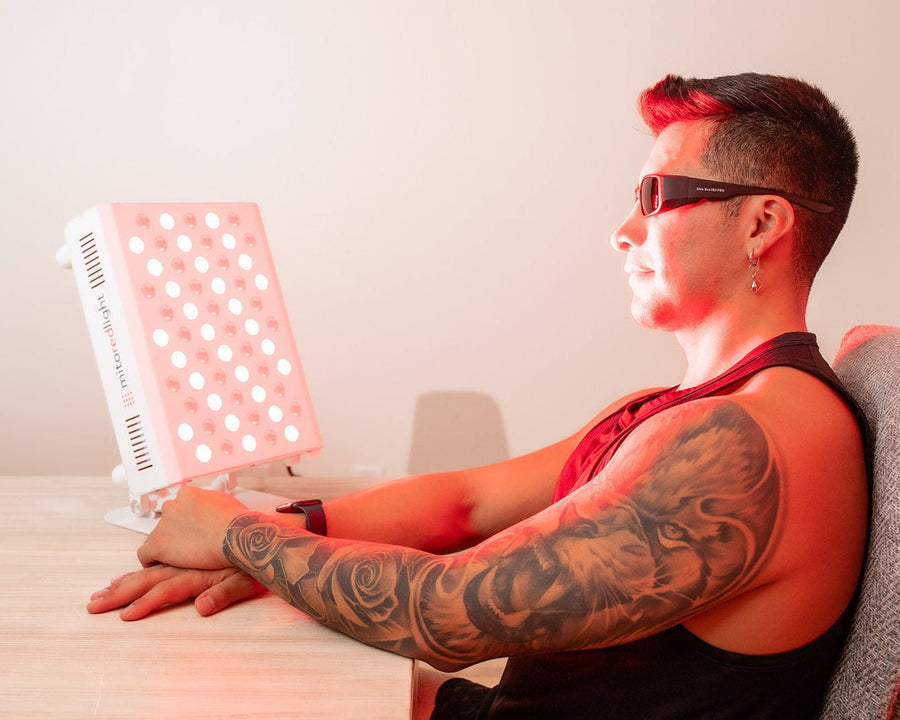 A person with sunglasses and a tattooed arm sits at a table using the MitoPRO+ Red Light Therapy Panel Series v2 by Mito Red Light, which emits bright red light onto their face and upper body with its quad wavelength design.