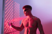 A shirtless man in sunglasses stands before the Mito Red Light MitoPRO+ | Red Light Therapy Panel Series v2, spraying mist on his face and illuminated by its pinkish-red light against a gradient backdrop.