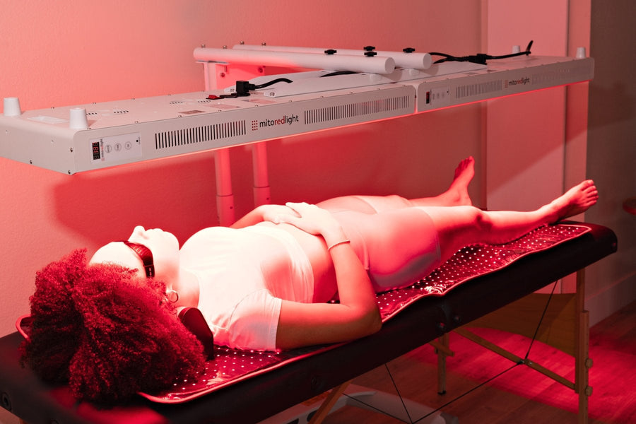 A person lies relaxed on a treatment table under Mito Red Light’s MitoPRO+ | Red Light Therapy Panel Series v2 in a softly lit clinical setting, wearing protective eyewear and a white outfit with hands resting on their stomach.