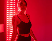 A woman in a black crop top stands before the MitoPRO+ | Red Light Therapy Panel Series v2 by Mito Red Light, bathed in red light. Tattoos adorn her arms as the quad wavelength design delivers optimal results.