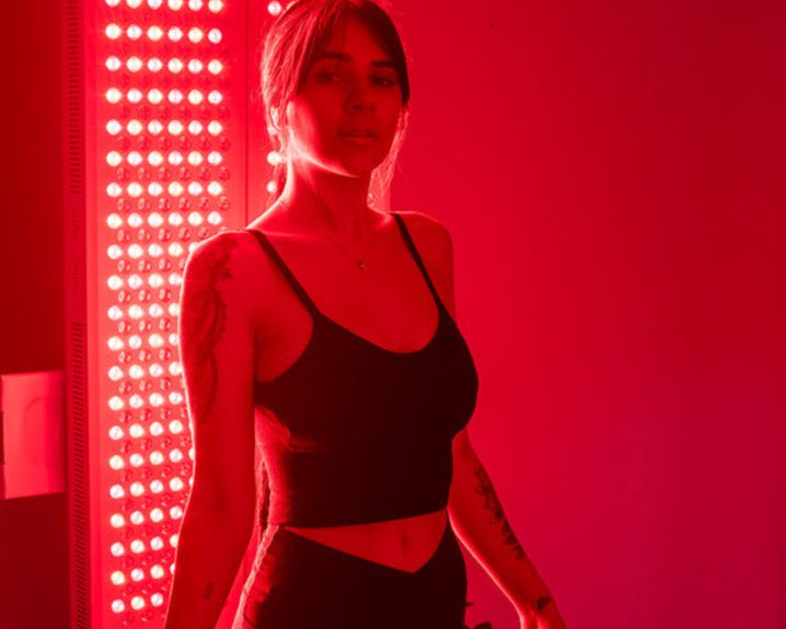 A woman in a black crop top stands before the MitoPRO+ | Red Light Therapy Panel Series v2 by Mito Red Light, bathed in red light. Tattoos adorn her arms as the quad wavelength design delivers optimal results.