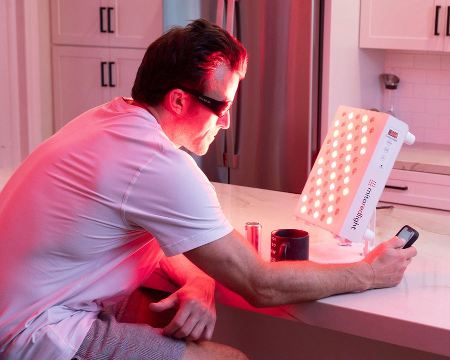 A man wearing sunglasses sits at a kitchen counter, using his phone beside a mug and can, all illuminated by the MitoPRO+ Red Light Therapy Panel Series v2 from Mito Red Light.