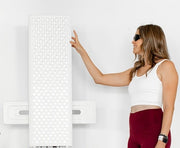 A woman in sunglasses, a white crop top, and maroon leggings stands next to the Mito Red Light MitoPRO+ | Red Light Therapy Panel Series v2, reaching out to touch it against a plain white background.