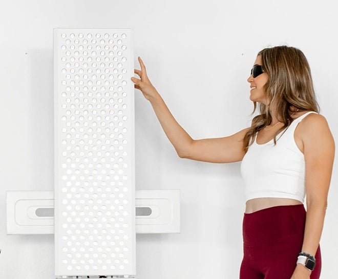 A woman in sunglasses, a white crop top, and maroon leggings stands next to the Mito Red Light MitoPRO+ | Red Light Therapy Panel Series v2, reaching out to touch it against a plain white background.