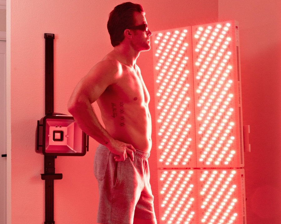 A shirtless man in sunglasses and gray shorts stands before a Mito Red Light MitoPRO+ Red Light Therapy Panel Series v2, bathed in red and near-infrared light in a pink-lit room.