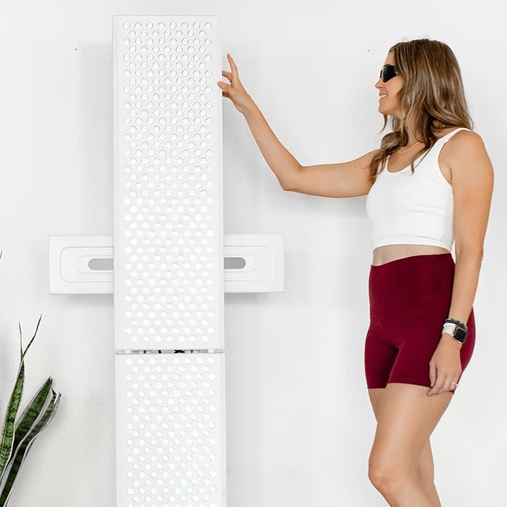 A woman in sunglasses, a white crop top, and burgundy shorts stands beside the tall MitoPRO+ | Red Light Therapy Panel Series v2 by Mito Red Light, touching its perforated surface. A plant is visible in the lower left against a white wall.