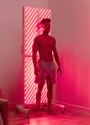 A shirtless man in shorts stands near a glowing MitoPRO+ | Red Light Therapy Panel Series v2 by Mito Red Light, bathing in red and near infrared light, in a sleek, modern room illuminated with a reddish-pink glow.